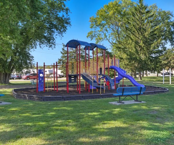 Colorful playground with slides and climbing bars, surrounded by trees, benches, and grassy area on a sunny day.